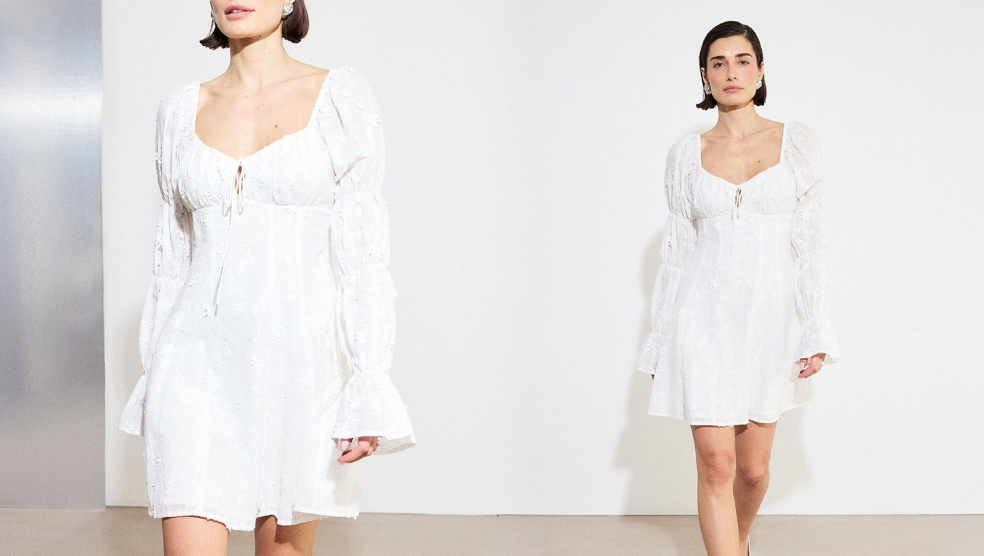 Model wearing a white graduation dress with long sleeves and tie-front detail, walking against a minimal studio background