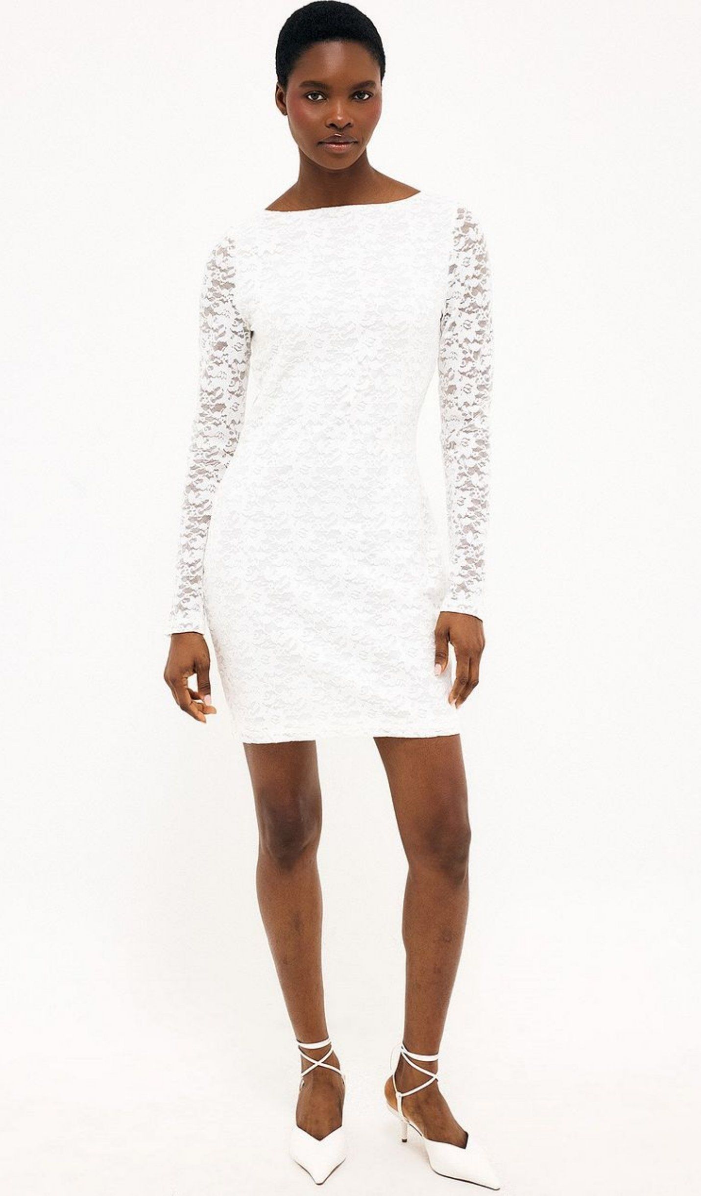 Model wearing a white lace mini dress with long sleeves and fitted silhouette, styled with strappy heels in a clean studio