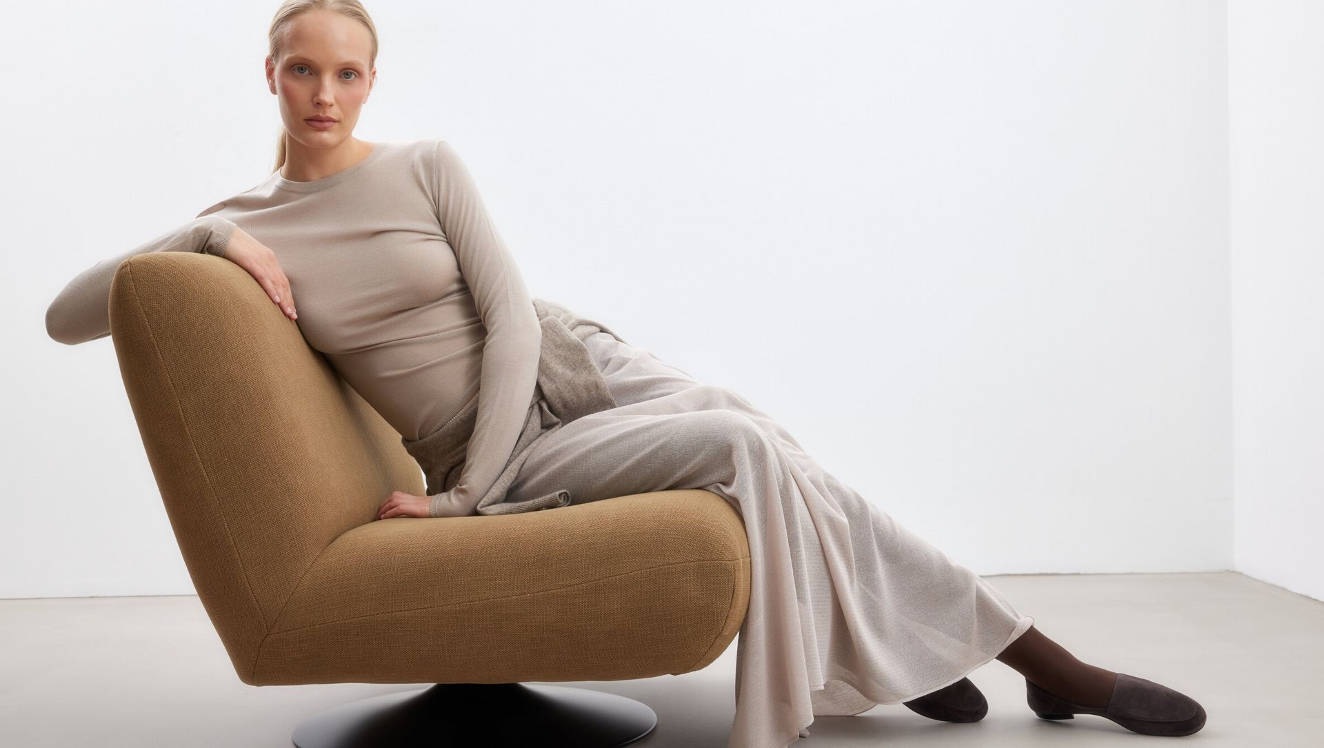 Model reclining on a mustard lounge chair wearing beige knit outfit and brown suede loafers