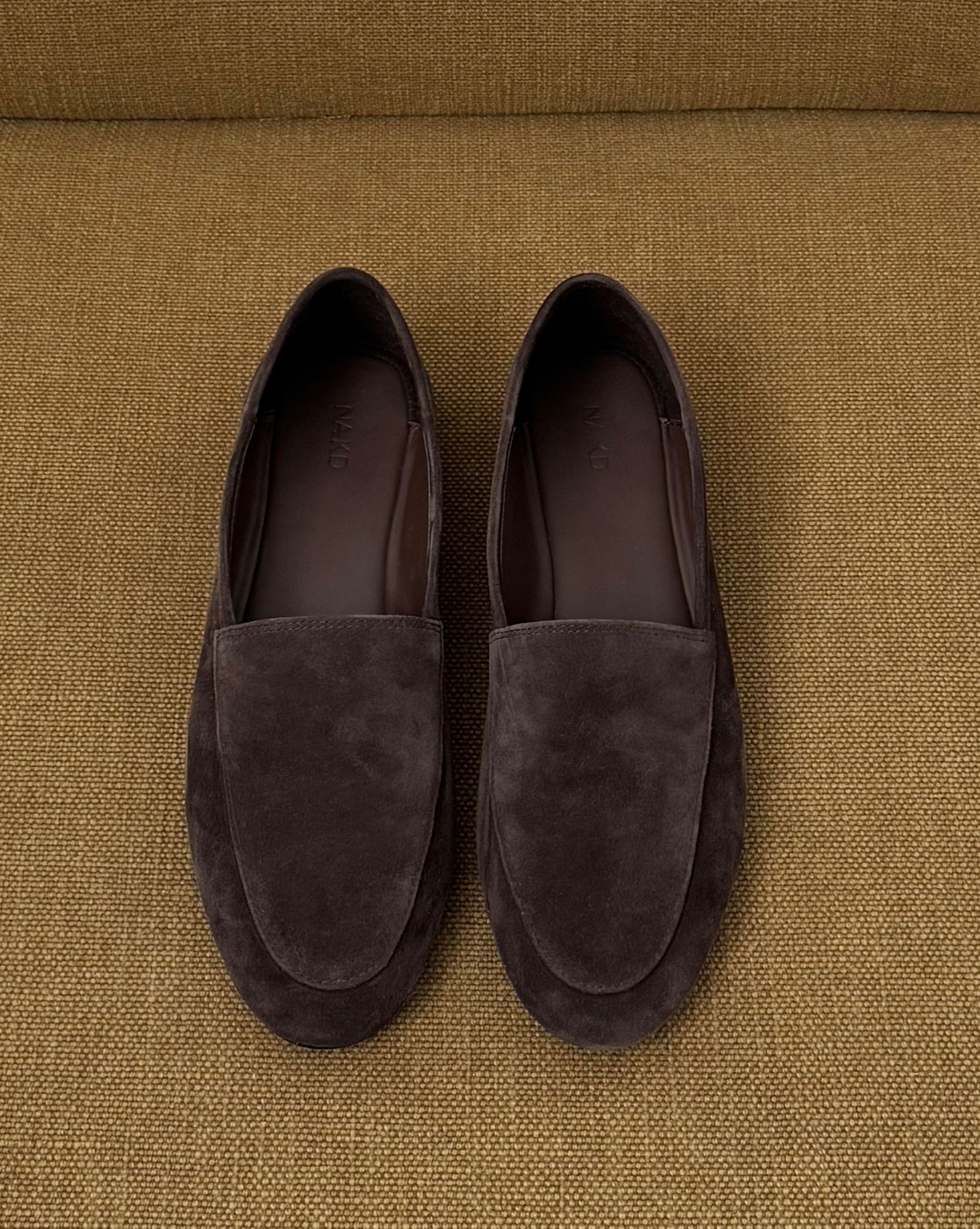 Pair of dark brown suede loafers displayed on textured mustard fabric background
