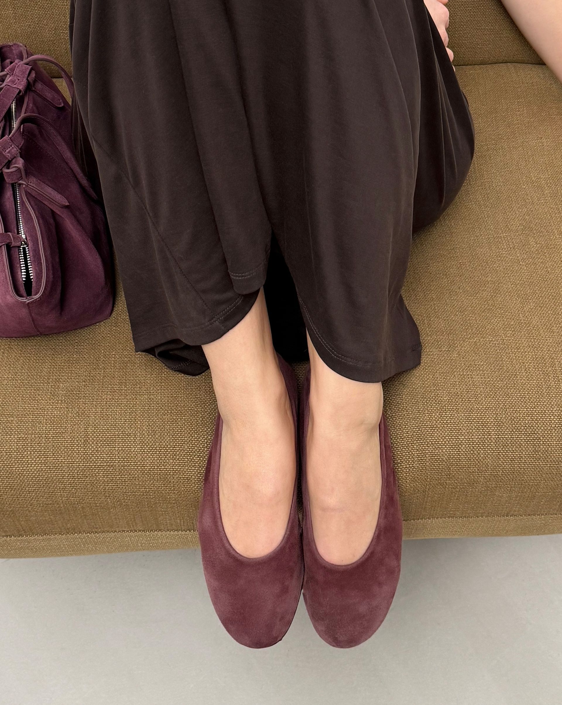 Detail of burgundy suede ballet flats worn with brown midi dress on lounge chair