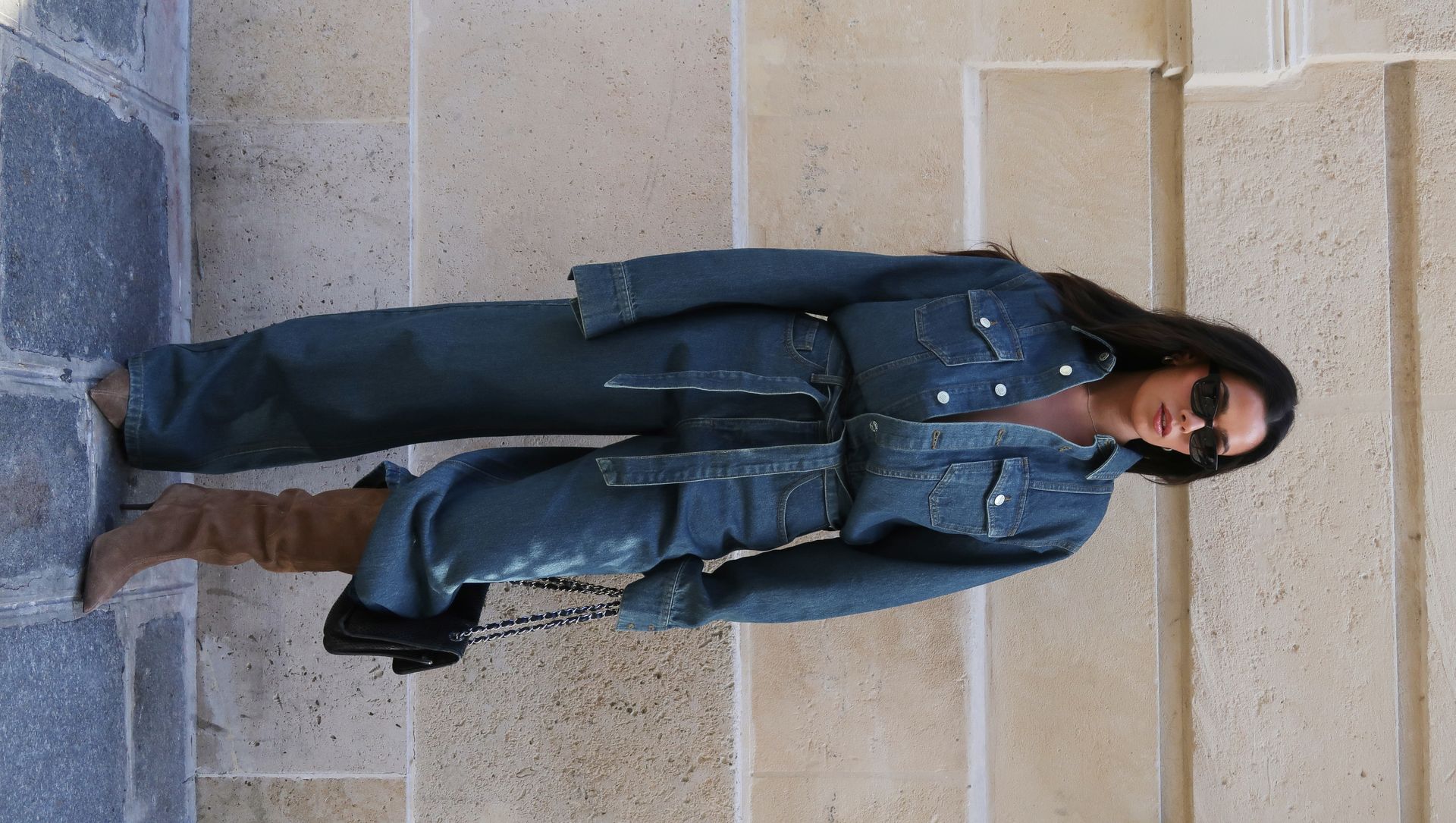 Woman in blue denim jumpsuit standing against stone wall wearing sunglasses and brown knee-high boots