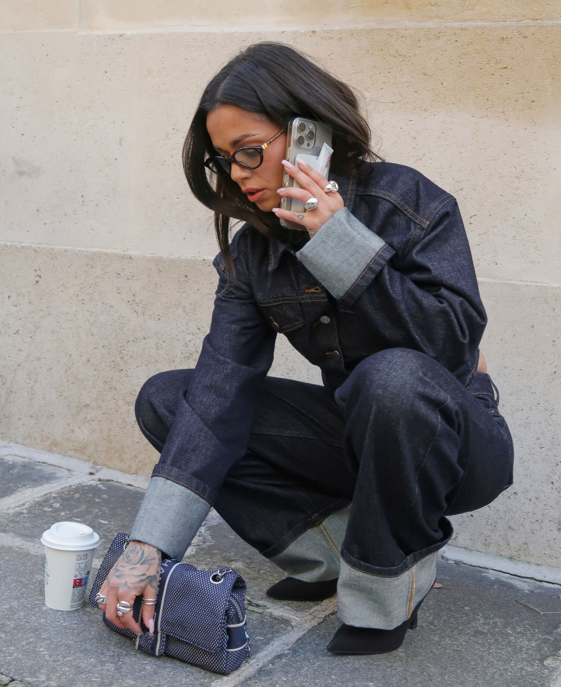 Woman in dark denim set crouching on sidewalk while talking on smartphone