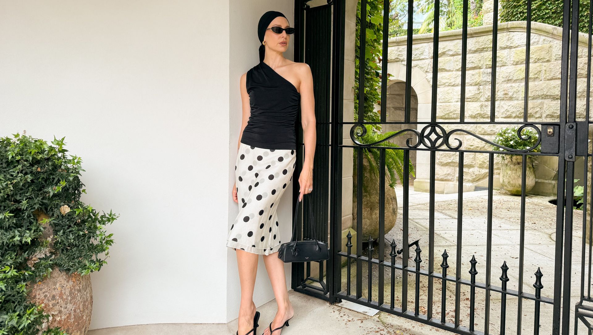 Kvinna i svart enaxlad topp och prickig kjol står vid en smidesgrind utomhus, med solglasögon, svart väska och sandaler.