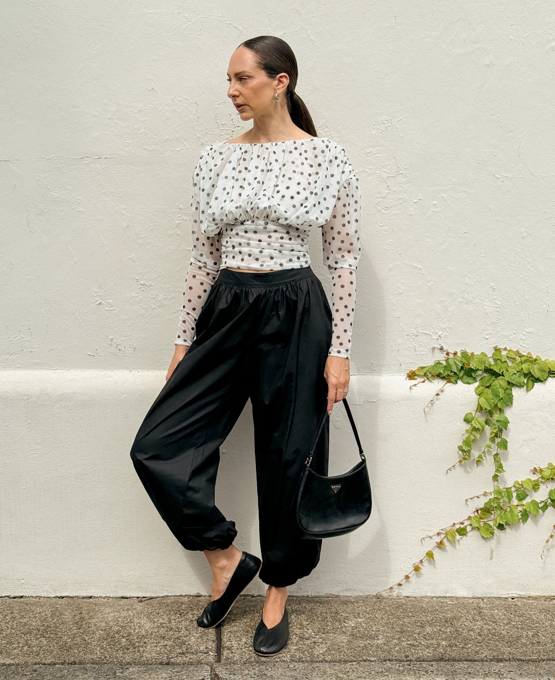 Kvinna i transparent prickig topp och svarta byxor lutar sig mot en ljus vägg, stylad med låg hästsvans och svart axelväska.