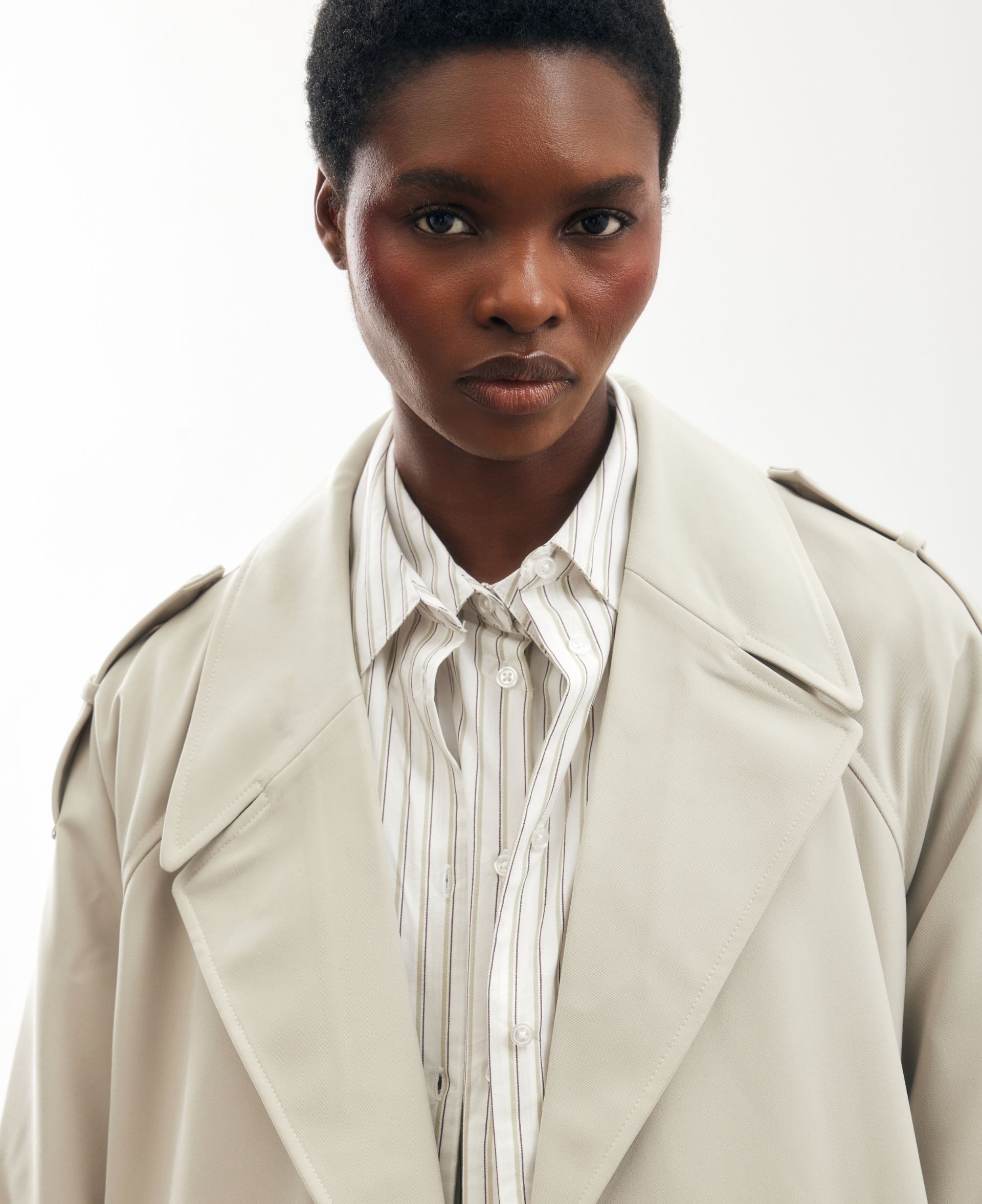 Model wearing a light beige trench coat layered over a striped button-up shirt, studio portrait on white background.