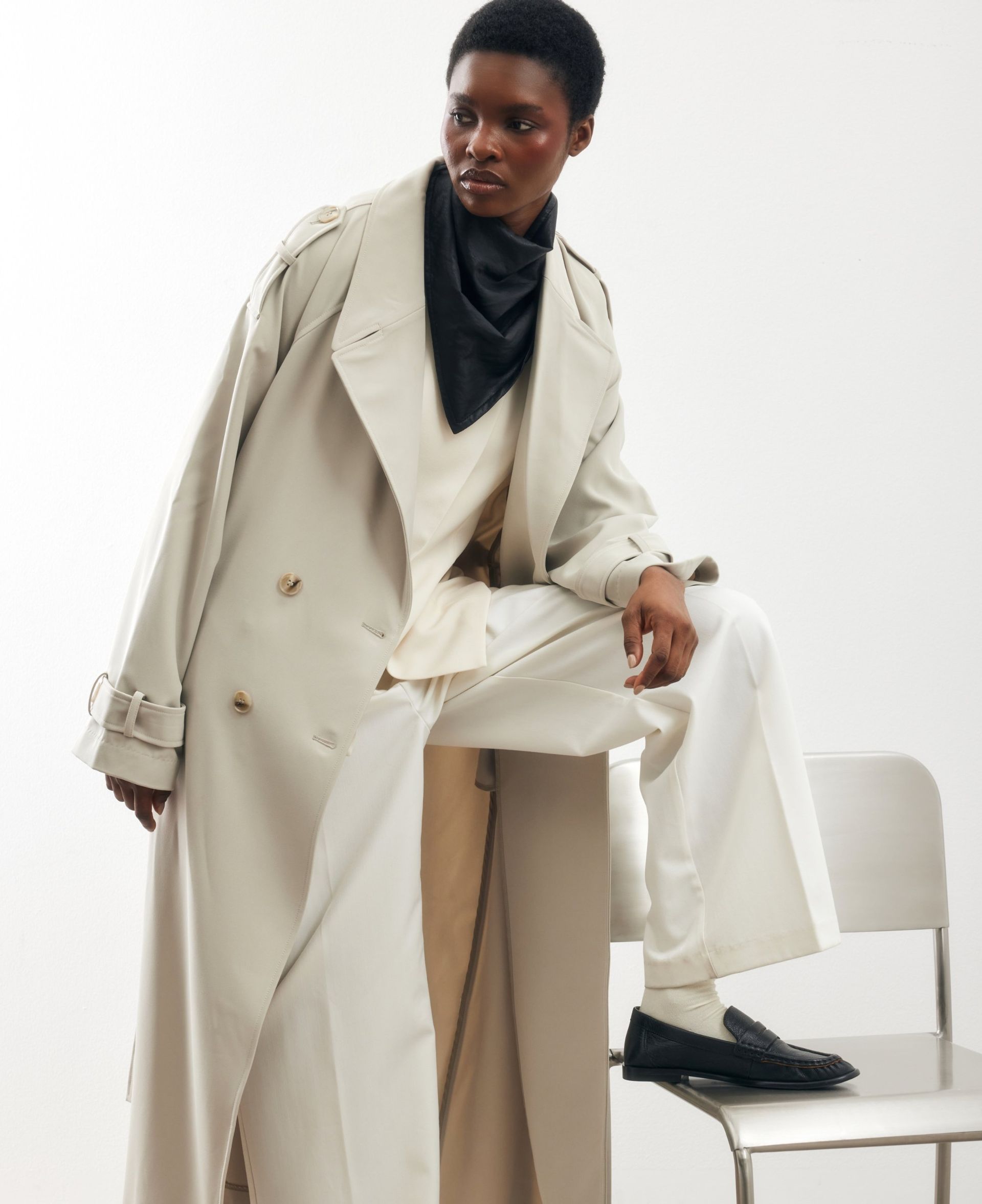 Model in oversized beige trench coat with cream trousers and black scarf, styled with loafers in a minimal studio setting.