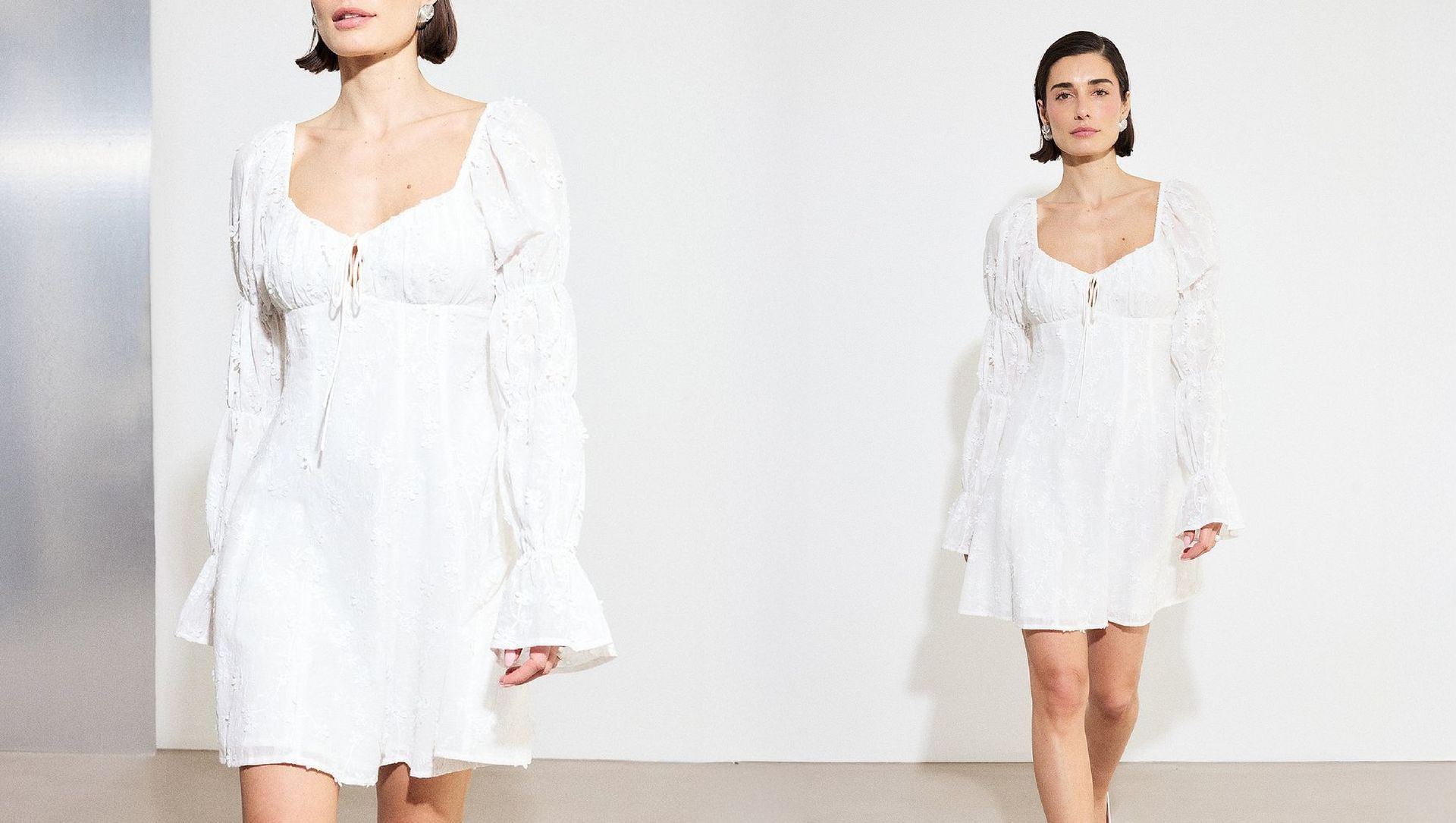 Model wearing a white graduation dress with long sleeves and tie-front detail, walking against a minimal studio background