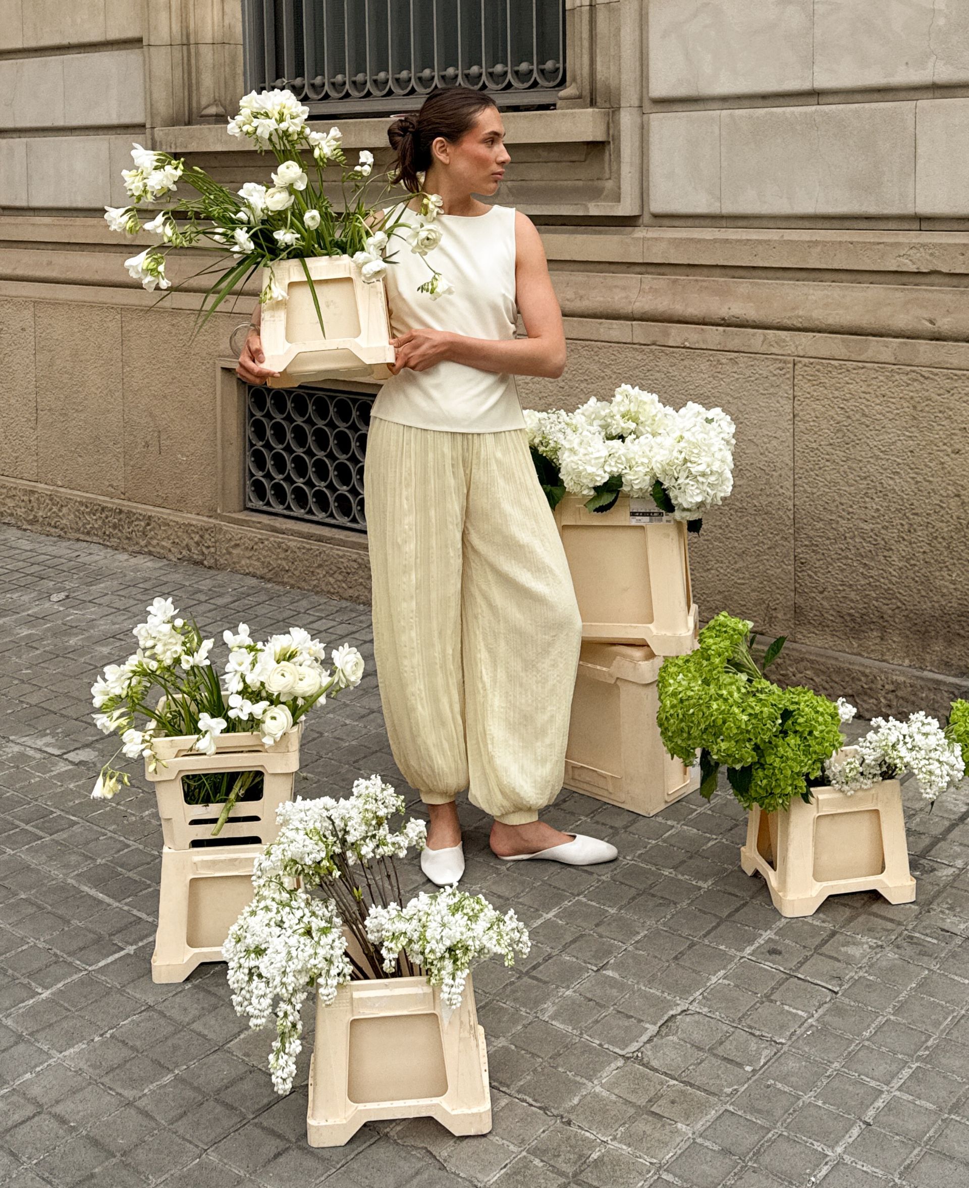 Woman in cream balloon pants and sleeveless top standing on sidewalk surrounded by flower bouquets against a stone building backdrop